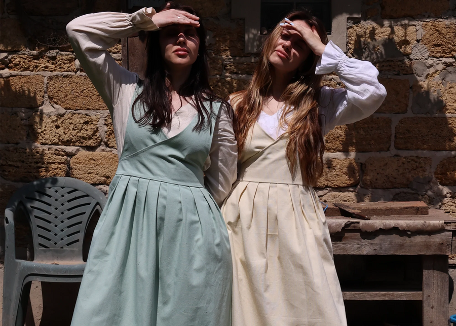 Two women wearing long white long-sleeve dresses with apron-style front panels, standing outdoors in front of a rustic stone wall. One dress is a pale sage green and the other is a warm cream/beige, both with full gathered skirts.