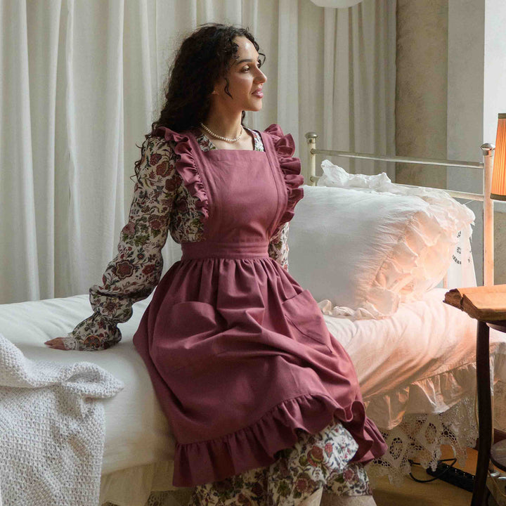 Woman in a vintage apron sitting on a bed in a softly lit room.