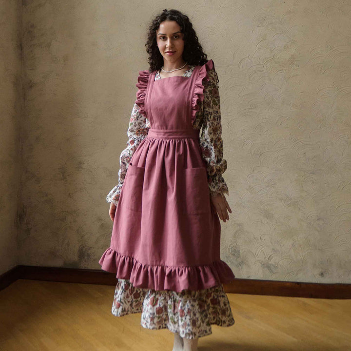 Woman wearing a pink apron over a floral dress against a beige wall.