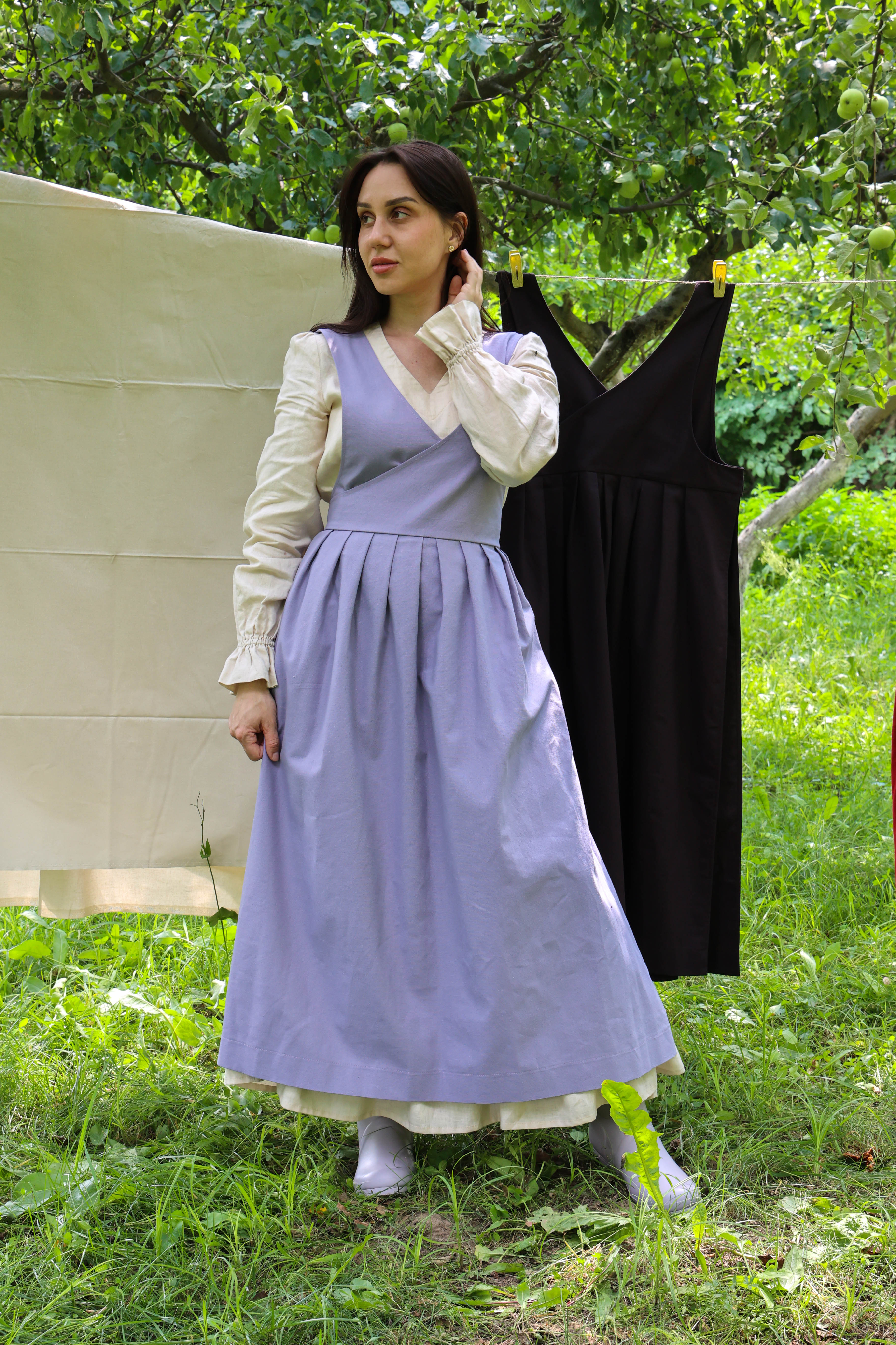 A woman stands outdoors in a grassy area wearing a long lavender apron over a white blouse, with a black dress hanging on a clothesline behind her and trees in the background.