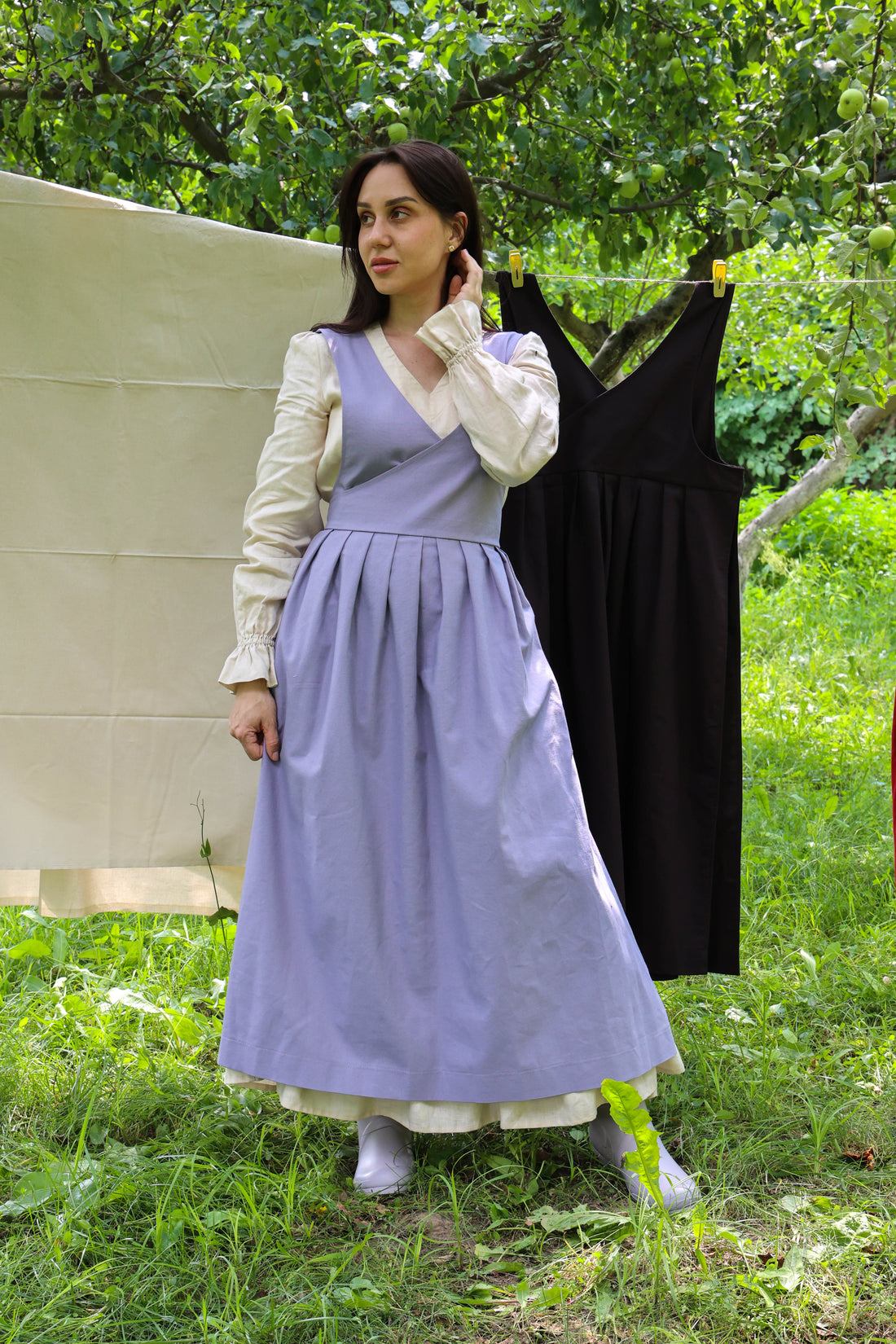 A woman stands outdoors in a grassy area wearing a long lavender apron over a white blouse, with a black dress hanging on a clothesline behind her and trees in the background.