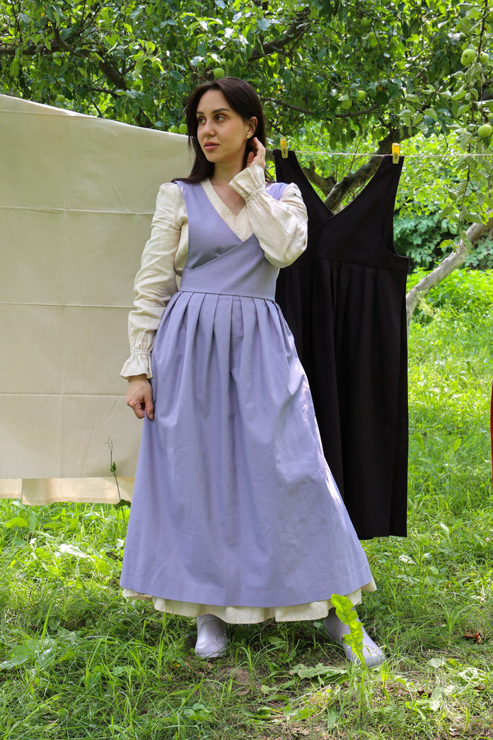A woman stands outdoors in a grassy area wearing a long lavender apron over a white blouse, with a black dress hanging on a clothesline behind her and trees in the background.