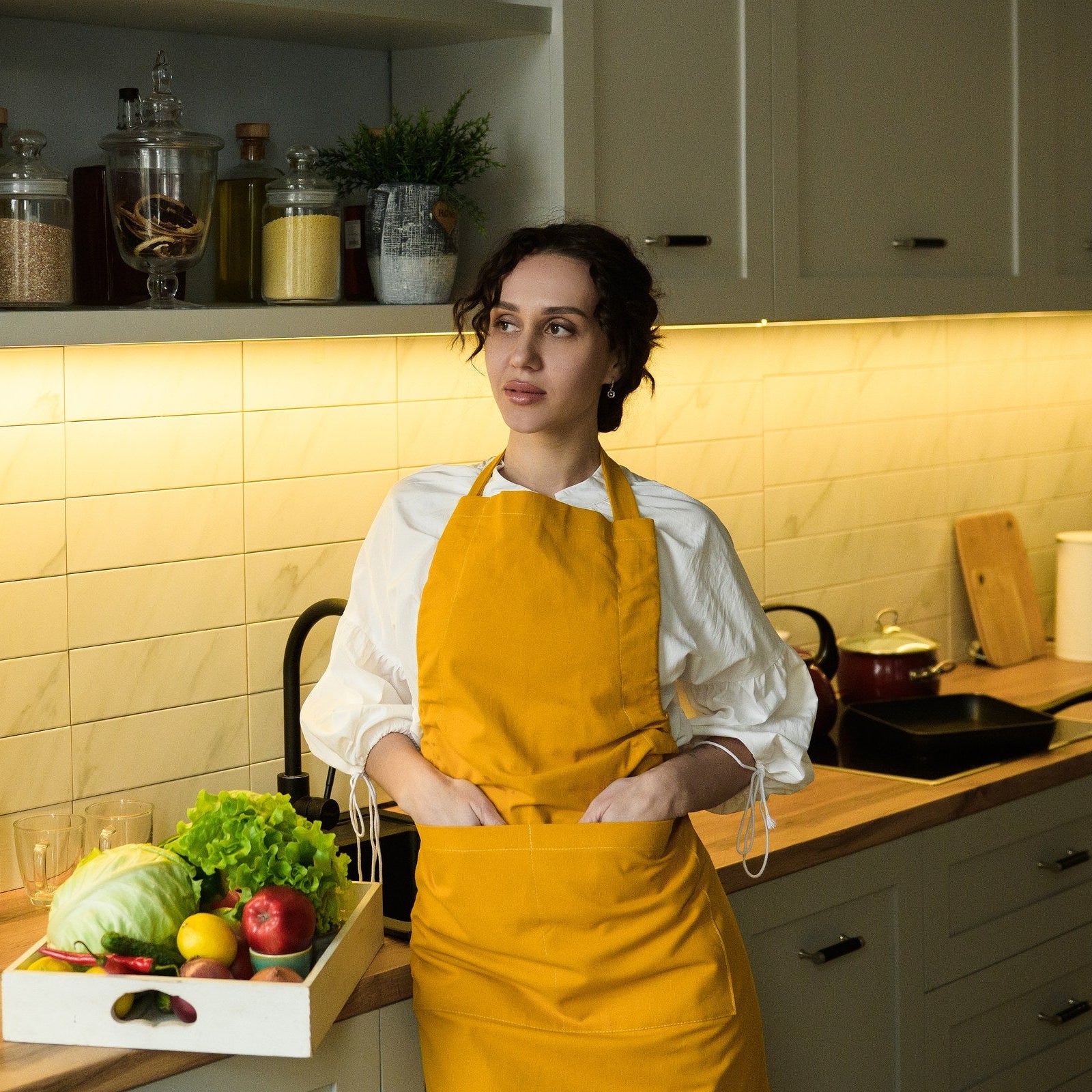Woman in a mustard yellow cotton bib apron standing at a kitchen counter