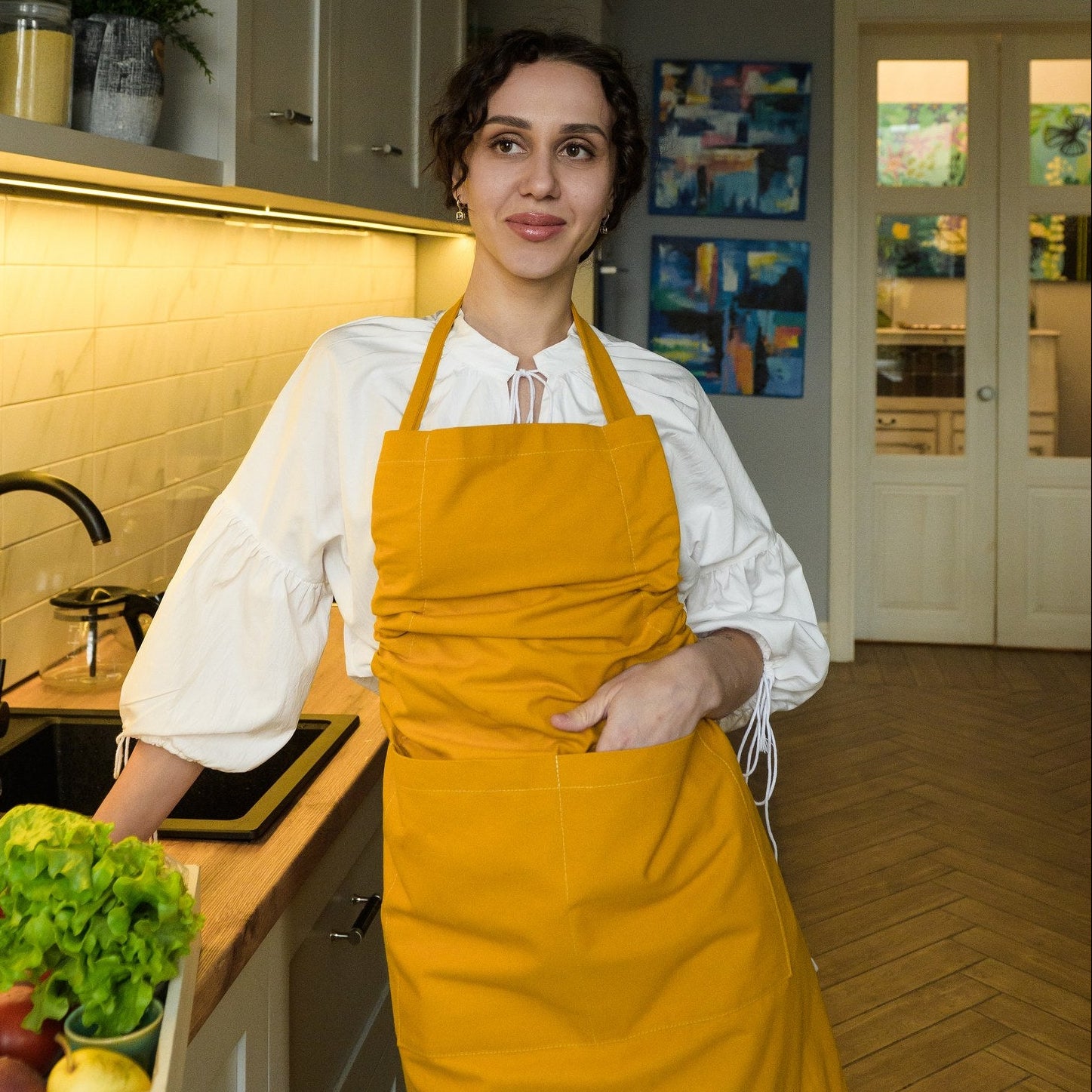 Mustard bib apron worn over a white blouse, front view with a hand in the pocket