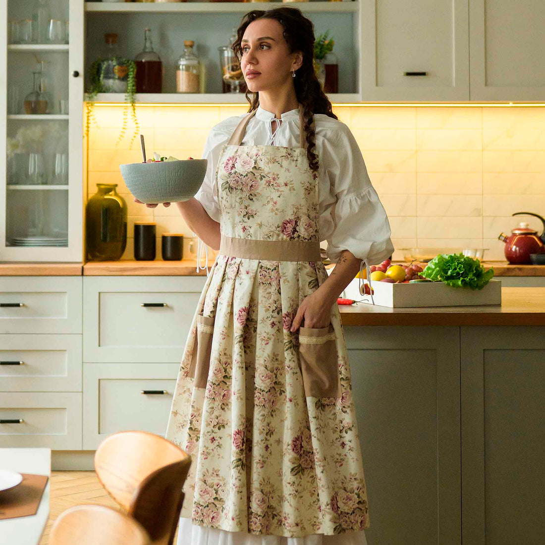 Woman in a kitchen wearing a floral apron holding a bowl.
