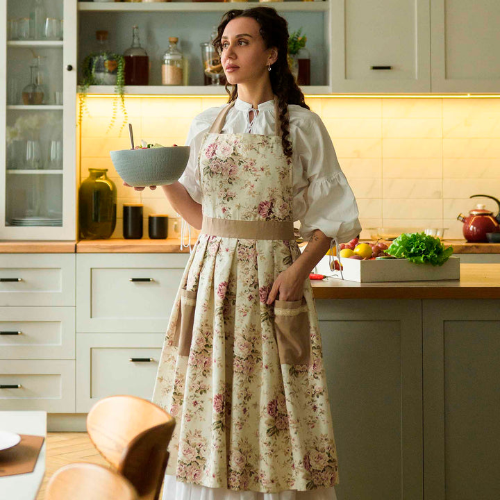 Woman in a kitchen wearing a floral apron holding a bowl.