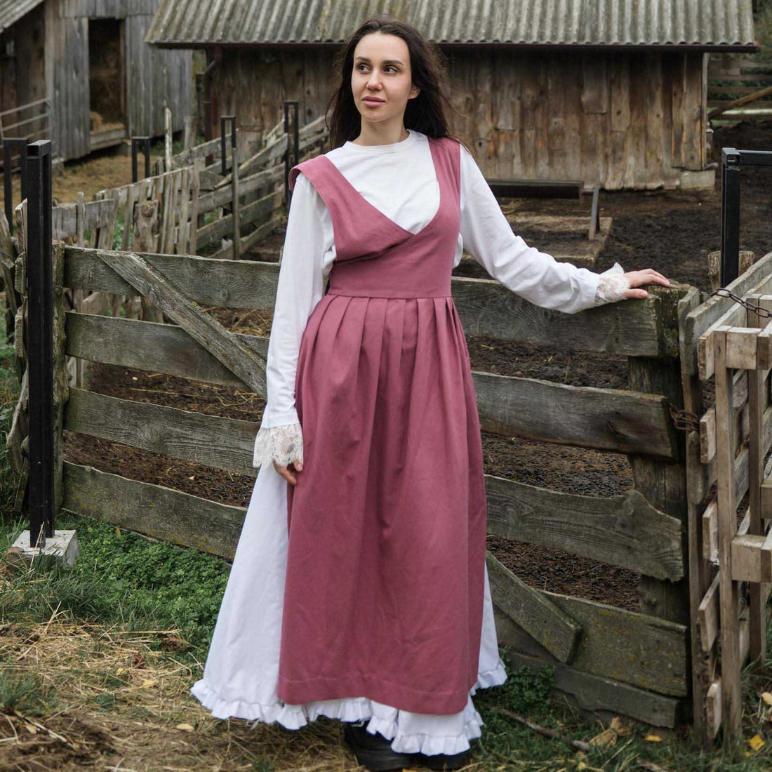 Woman in historical attire standing in front of wooden buildings and a fence.