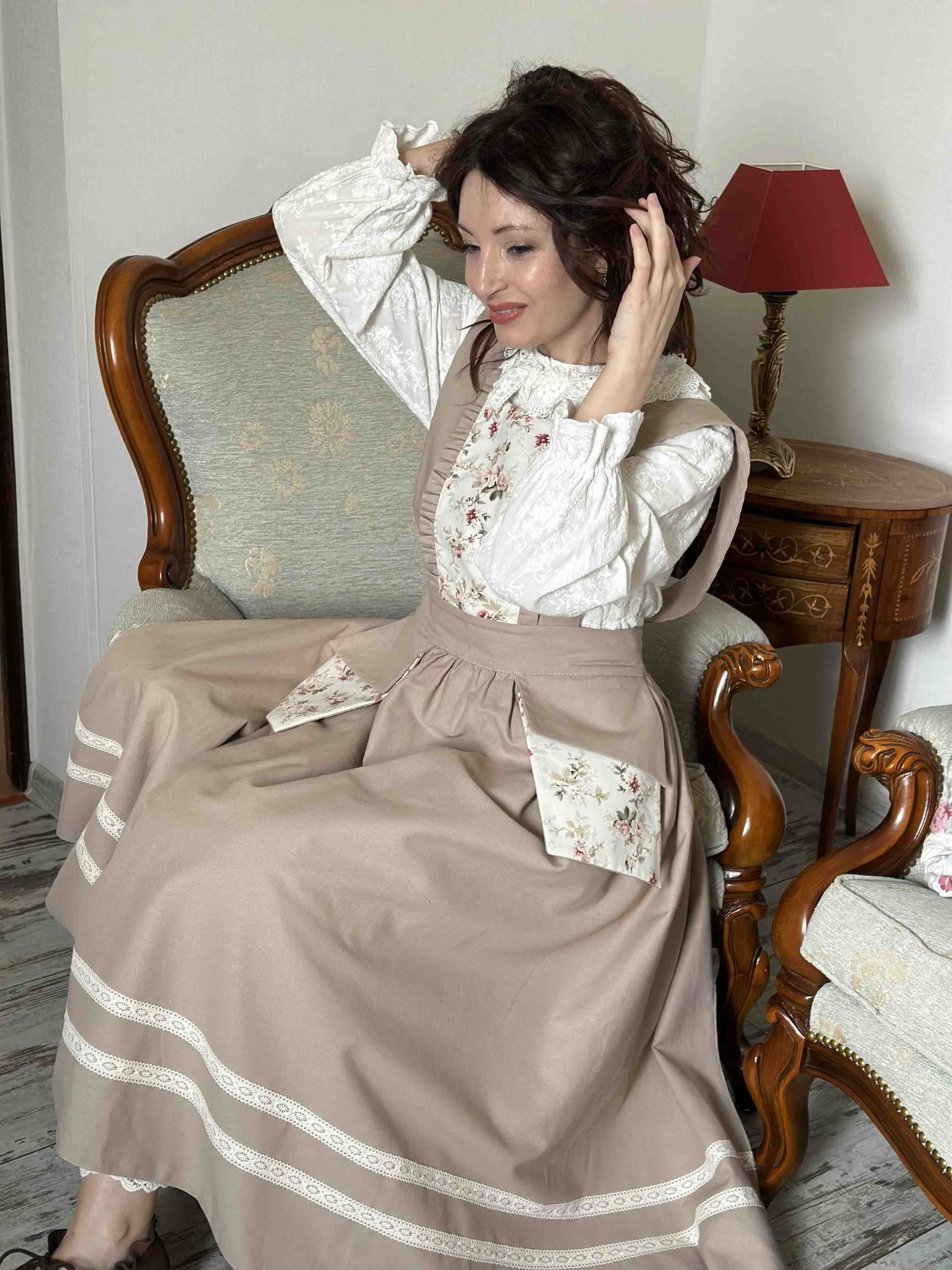 Woman in a vintage-style dress sitting on a wooden chair in a room with a lamp and cushion.