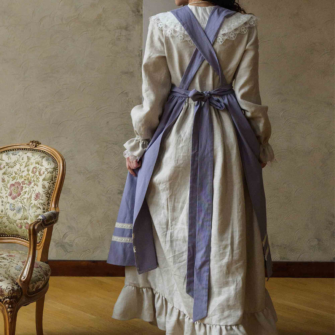 Person wearing a long beige dress with a purple apron in a room with a wooden chair and patterned wallpaper.