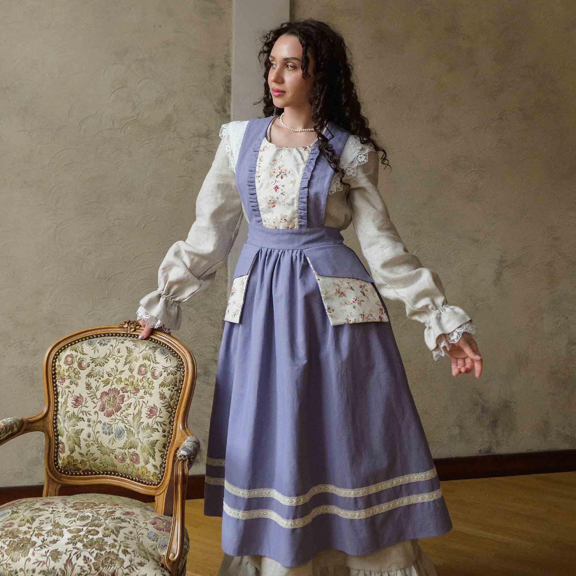 Woman in a historical dress standing next to an ornate chair in a room with a textured wall.