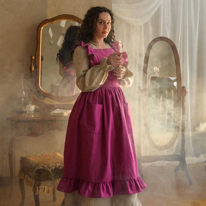 Woman in a pink apron holding a candle in a vintage room with mirrors and soft lighting.