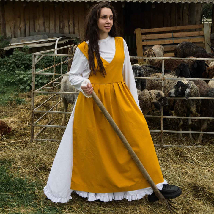 Woman in a yellow pinafore apron with a white shirt standing in a barn with sheep.
