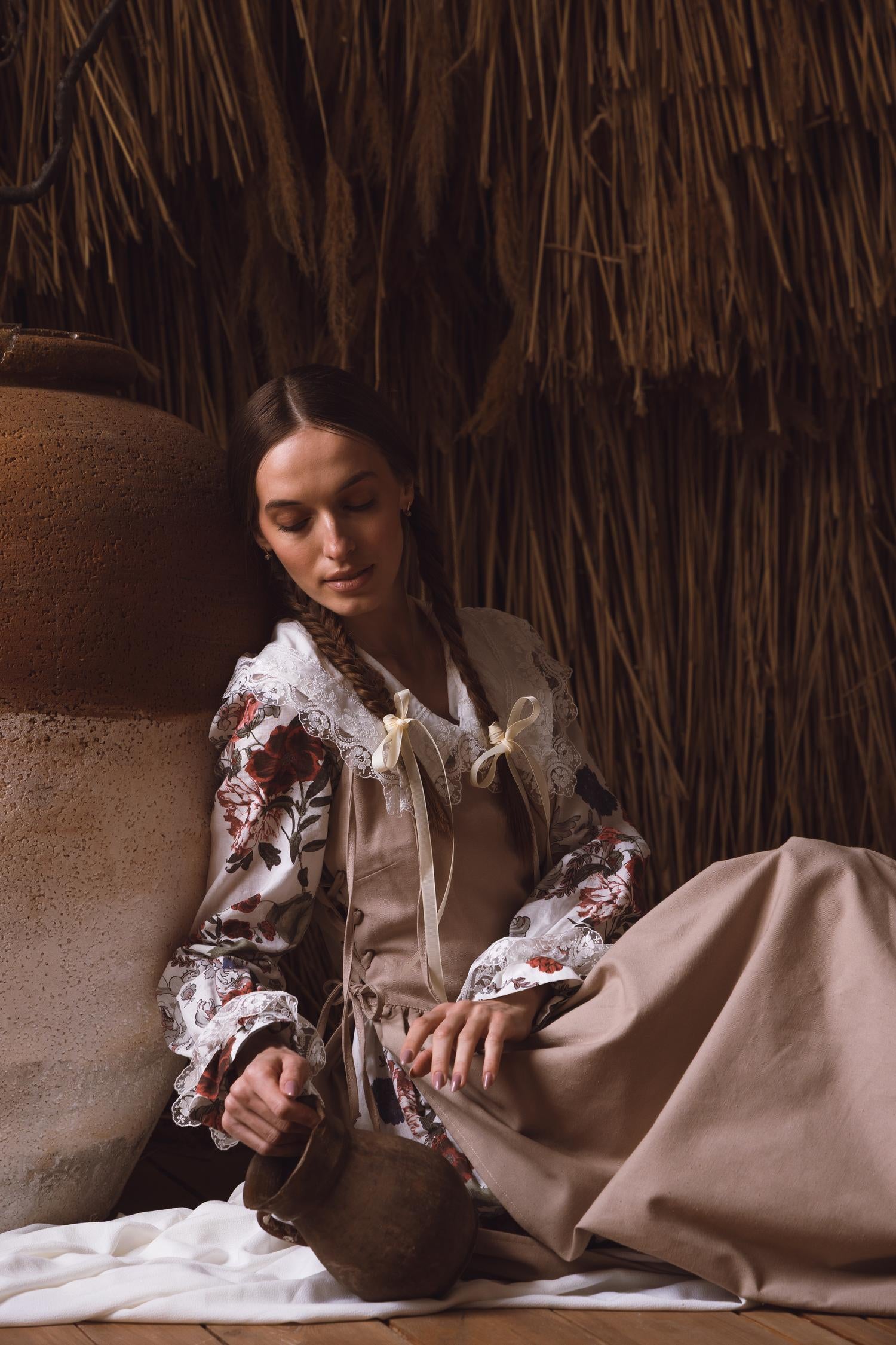 Woman in a floral blouse and beige skirt sitting against a thatched wall.