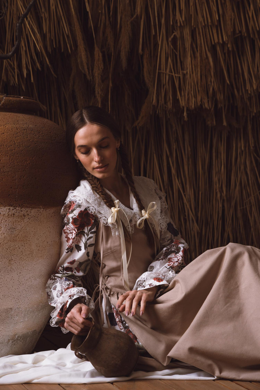 Woman in a floral blouse and beige skirt sitting against a thatched wall.