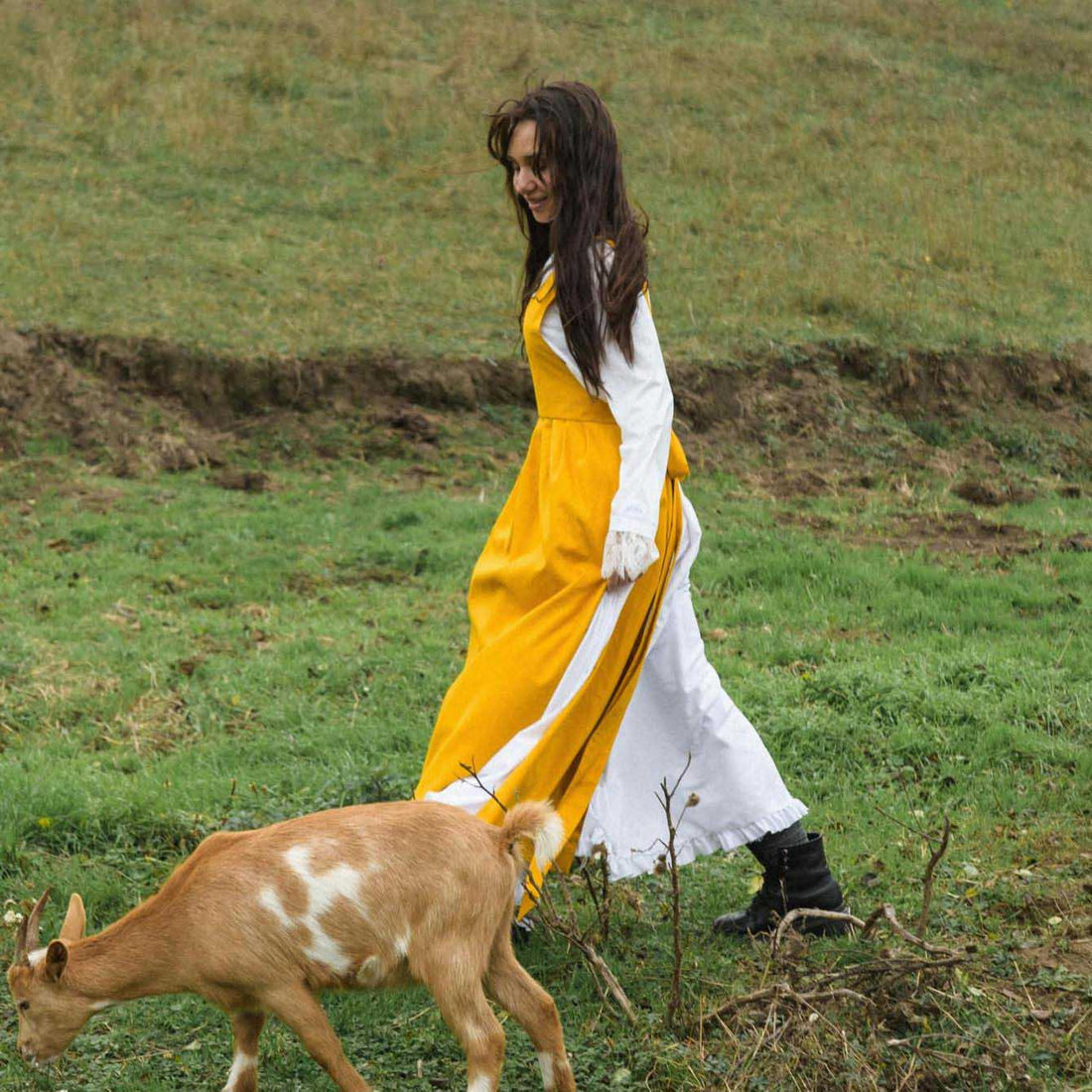 Woman in a yellow and white dress walking with a goat in a grassy field