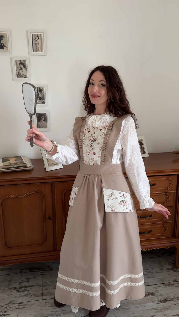 Woman in a vintage-style dress holding a fan in a room with wooden furniture and framed pictures on the wall.