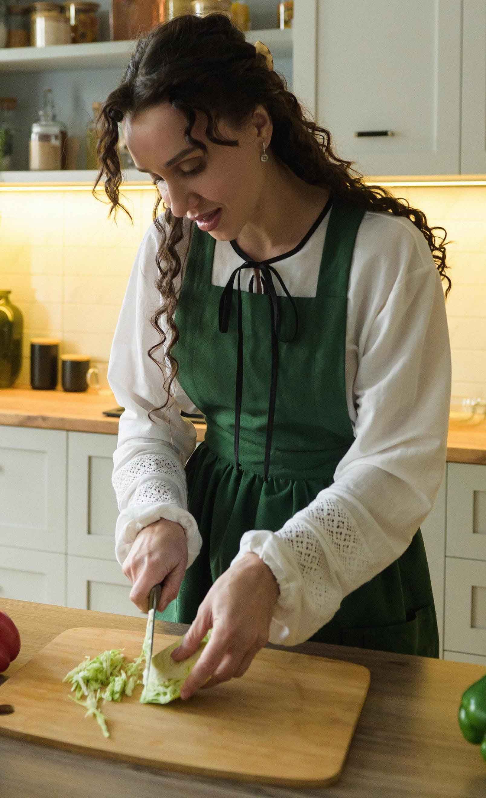 Woman in a green apron cutting vegetables on a wooden cutting board in a kitchen.