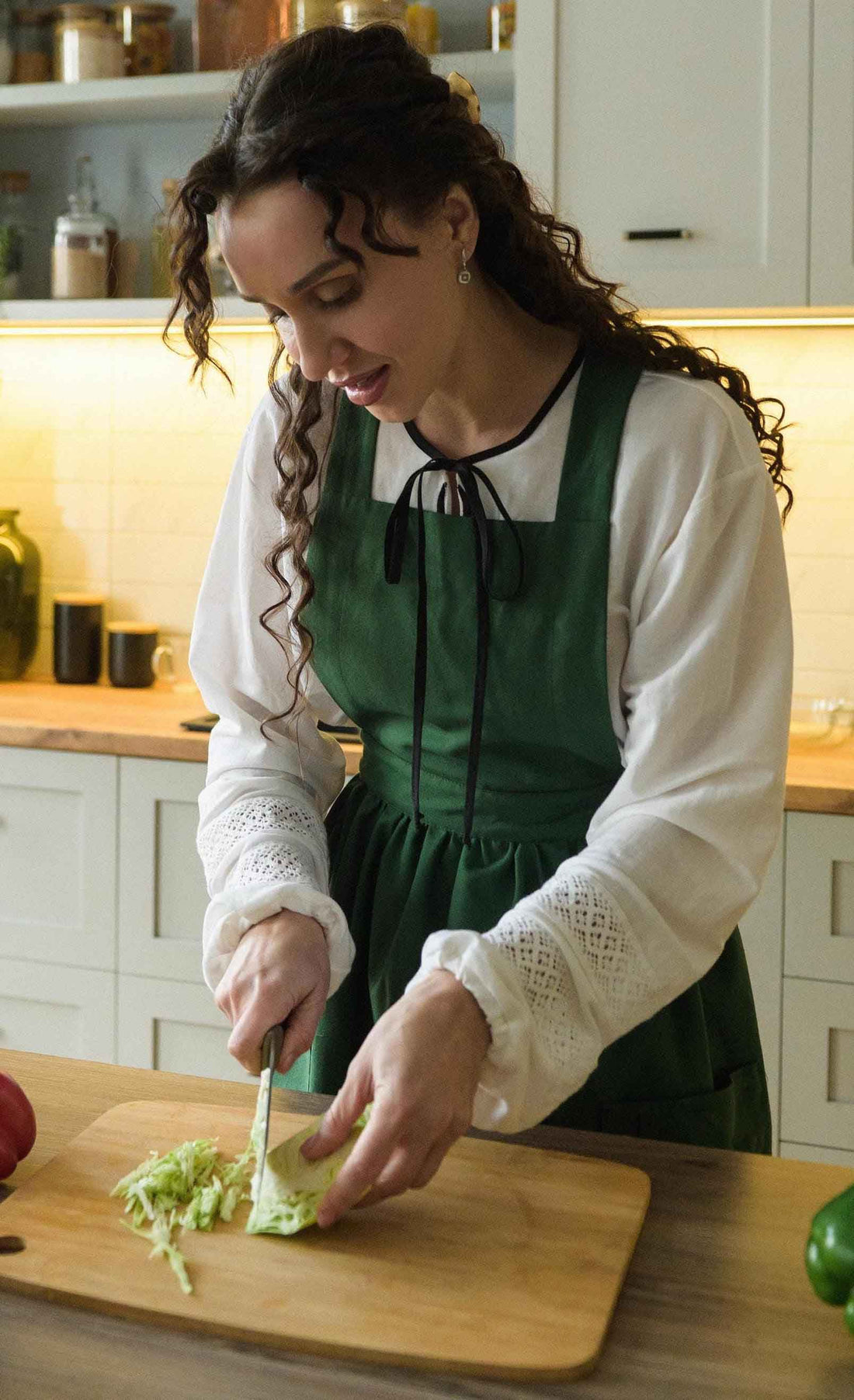 Woman in a green apron cutting vegetables on a wooden cutting board in a kitchen.