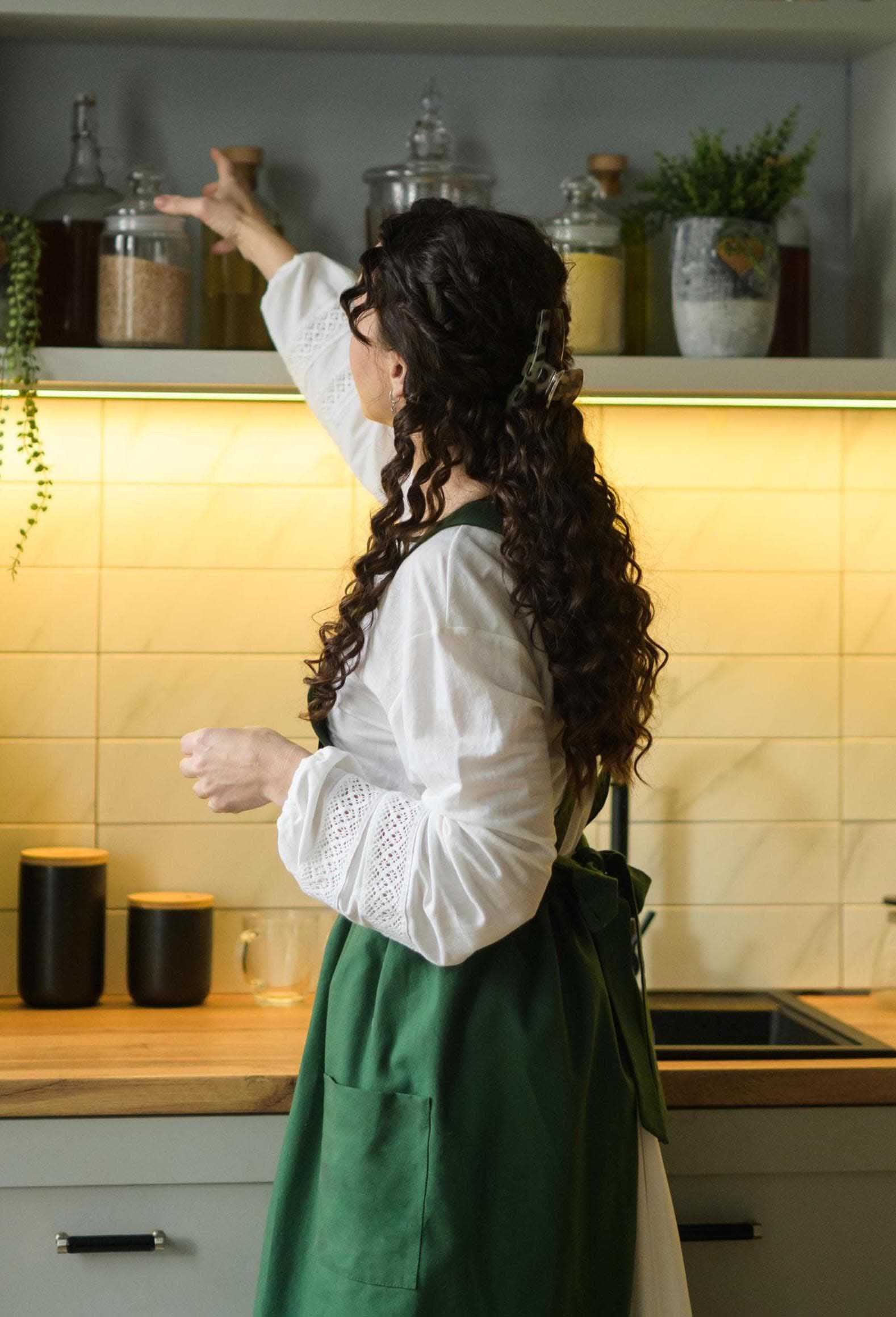 Woman in a kitchen wearing a white blouse and green apron.