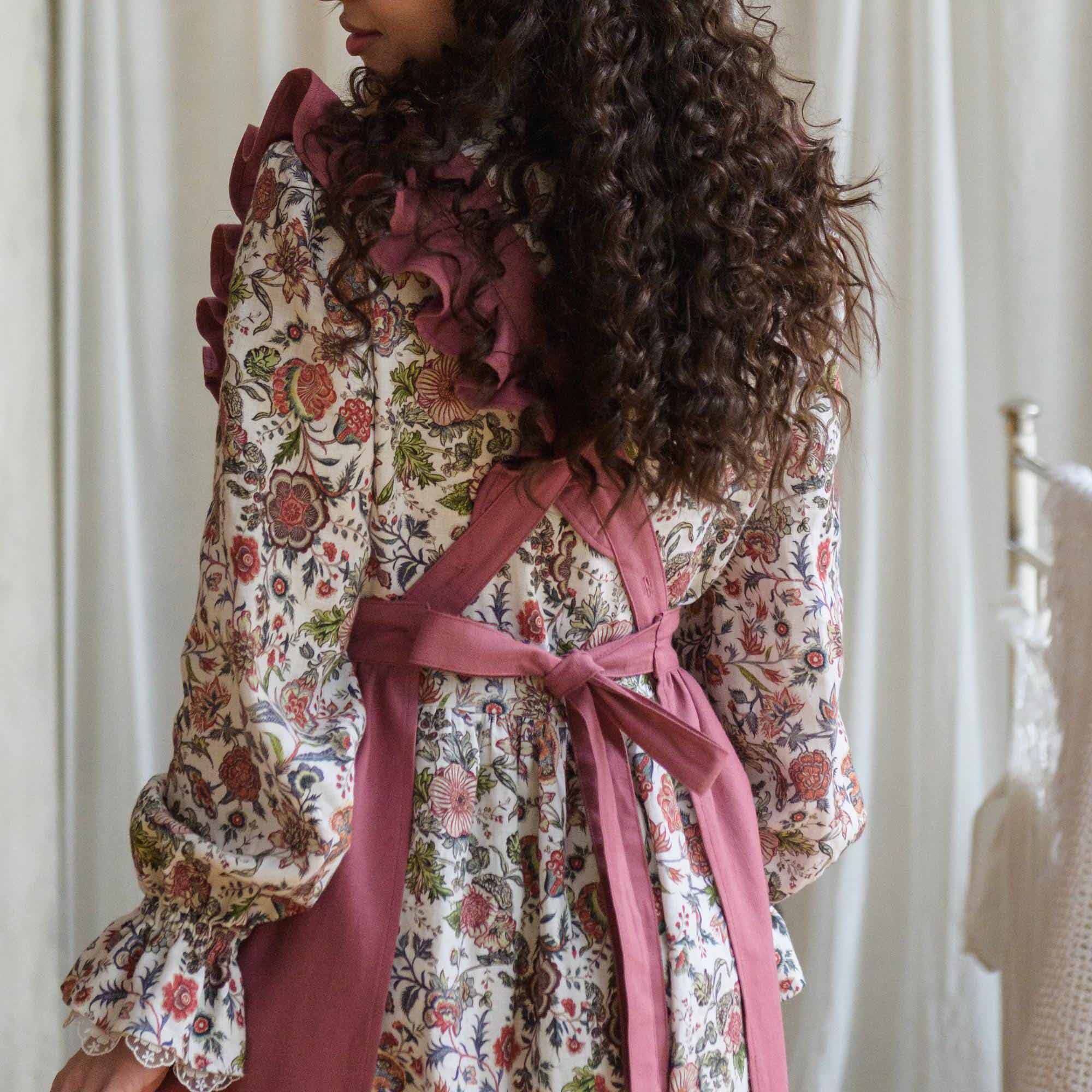 Woman wearing a pink apron with ruffled sleeves and a pink belt against a white curtain background