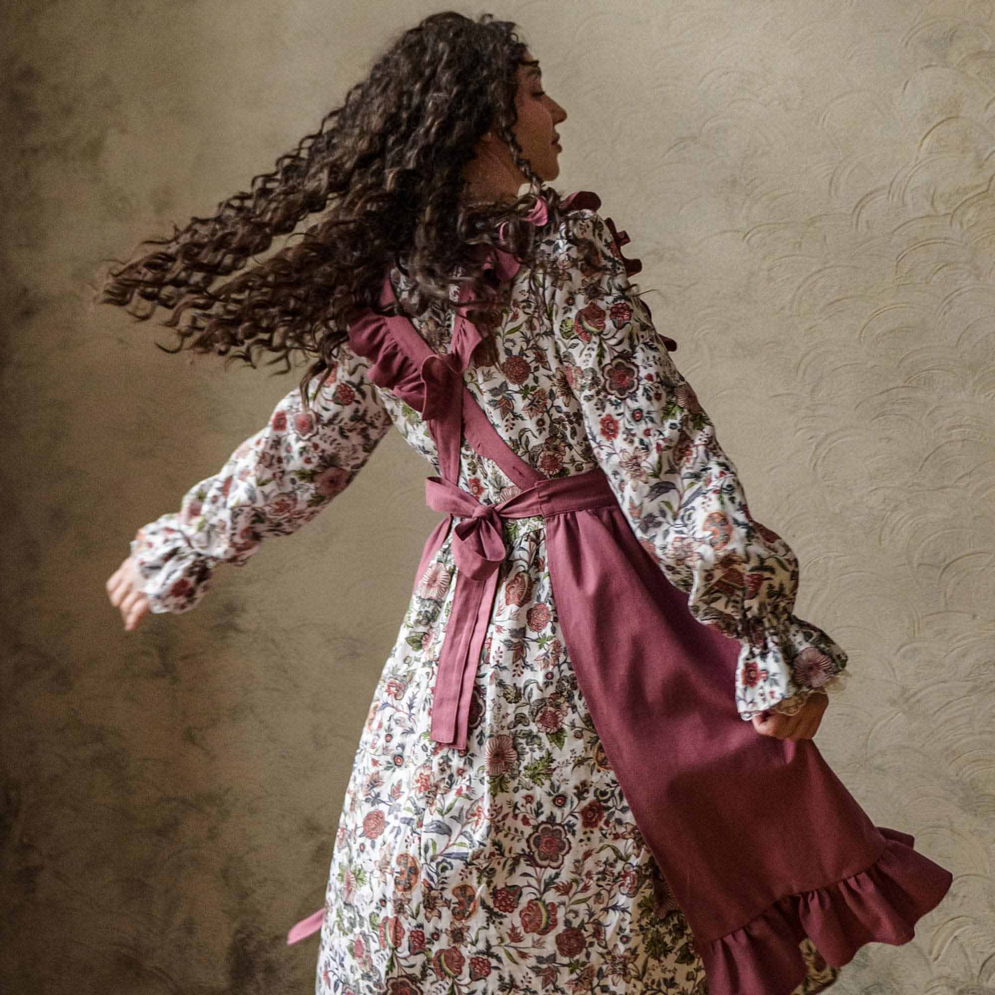 Woman in a pink apron with a burgundy belt against a textured beige wallk