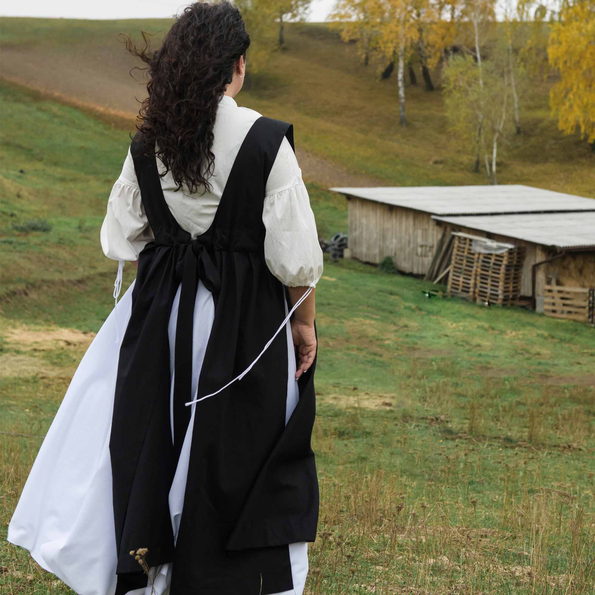 Person wearing a black apron over a white shirt in a rural setting with a barn and trees.