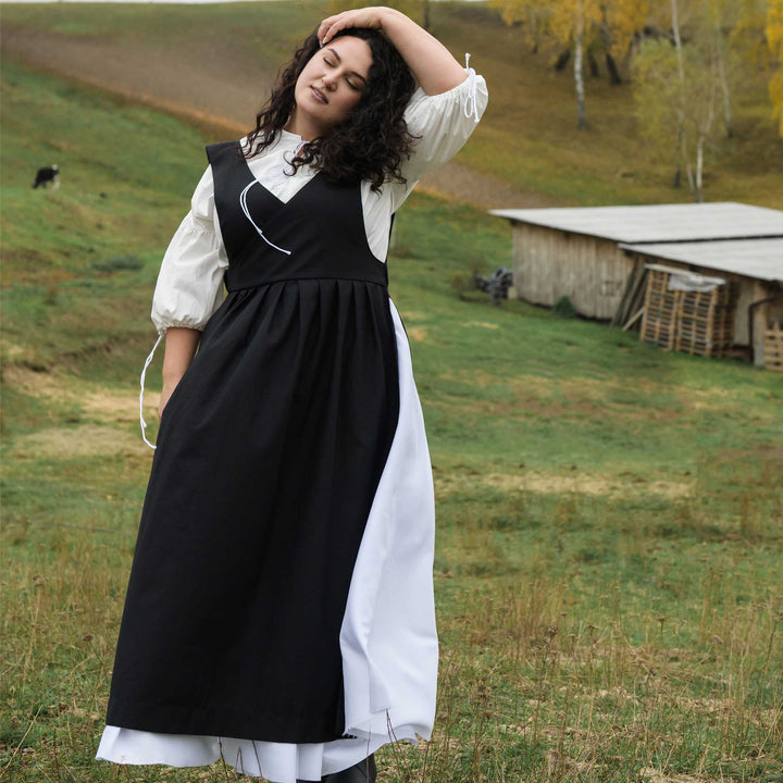 Woman in a black and white dress standing in a grassy field with a wooden cabin in the background.