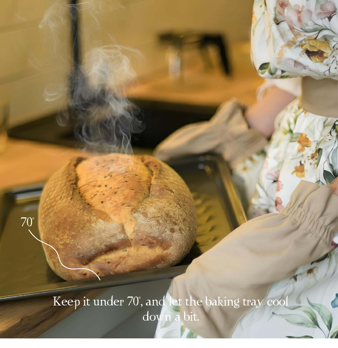 Loaf of bread on a baking tray with steam rising, person wearing floral apron in background