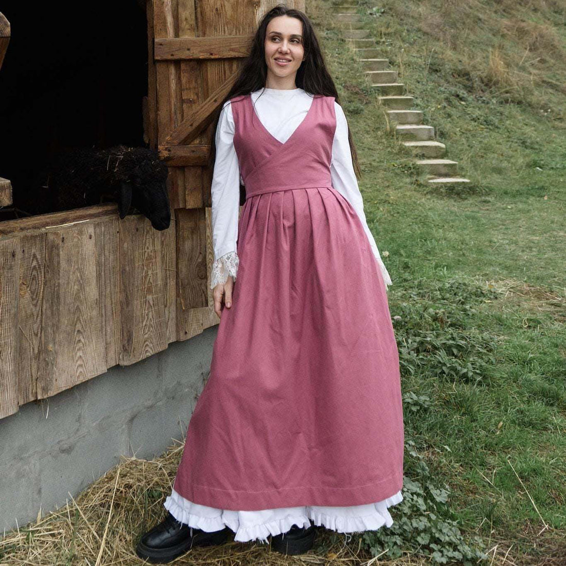 Woman in a pink apron pinafore standing in front of a wooden building with a horse.