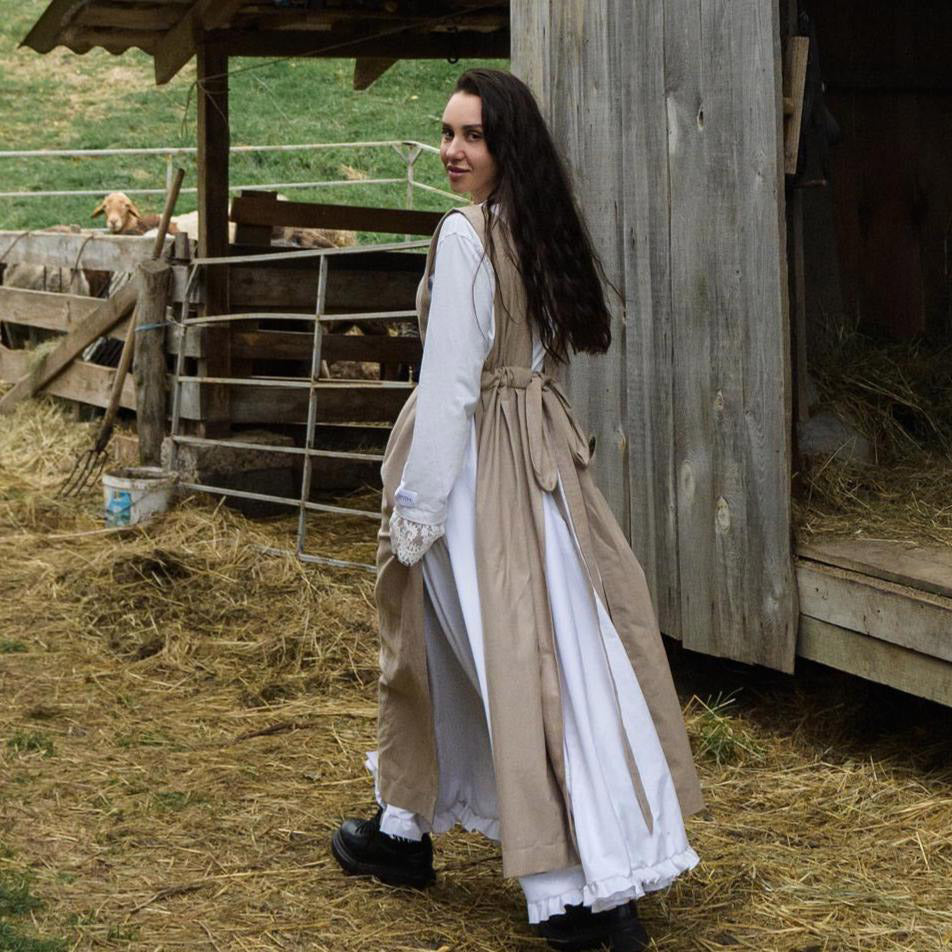 Woman in historical attire standing in a barn with hay and wooden structures.