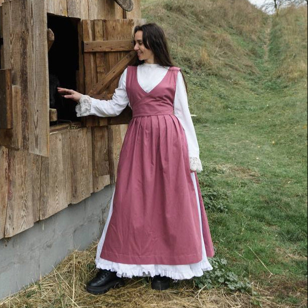 Woman in a pink pinafore apron standing next to a wooden structure outdoors.