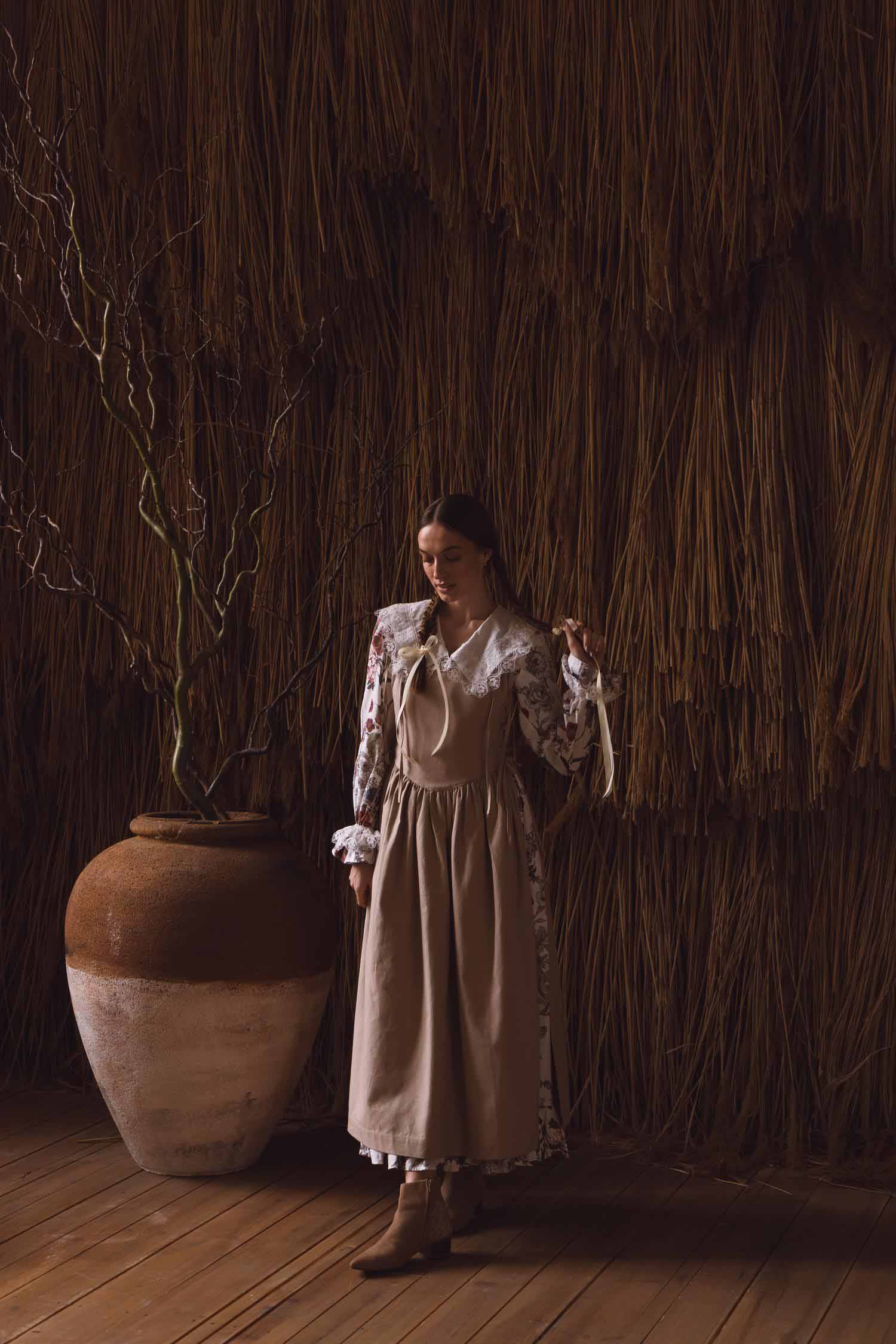 Woman in a vintage dress standing next to a large vase against a thatched wall.