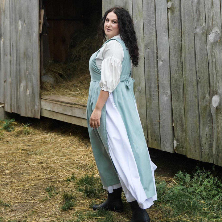 Woman in a light blue and white dress standing in front of a wooden structure with hay.