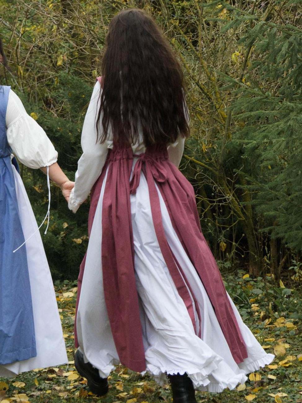 Two people in historical or period costumes walking outdoors with greenery in the background.